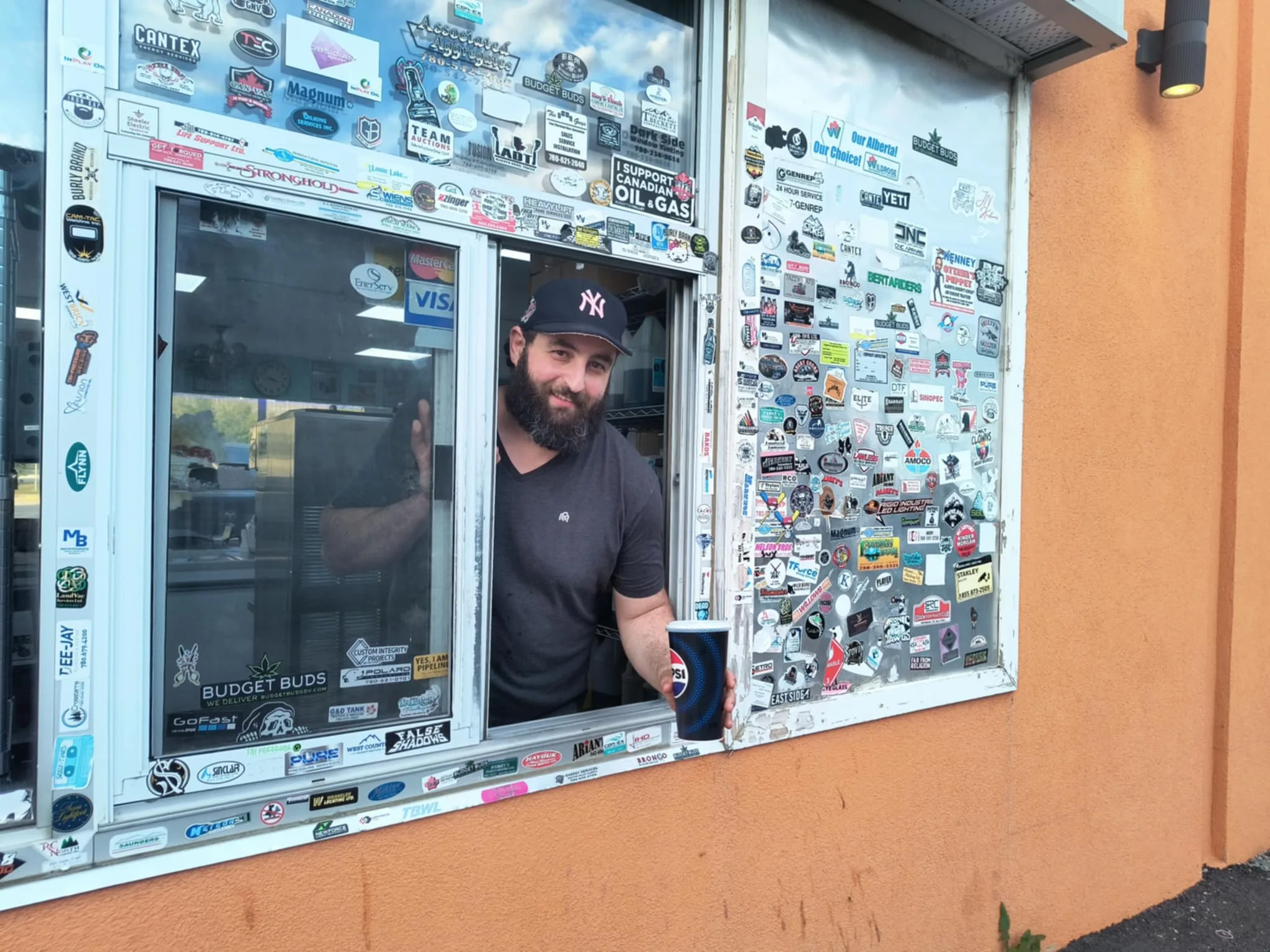 Sticker Wall Drive-Thru – Burger Baron Drayton Valley Close-up image of the sticker-covered drive-thru window at Burger Baron in Drayton Valley, Alberta, which was reimagined in the AI video as a cave art-covered prehistoric ordering window